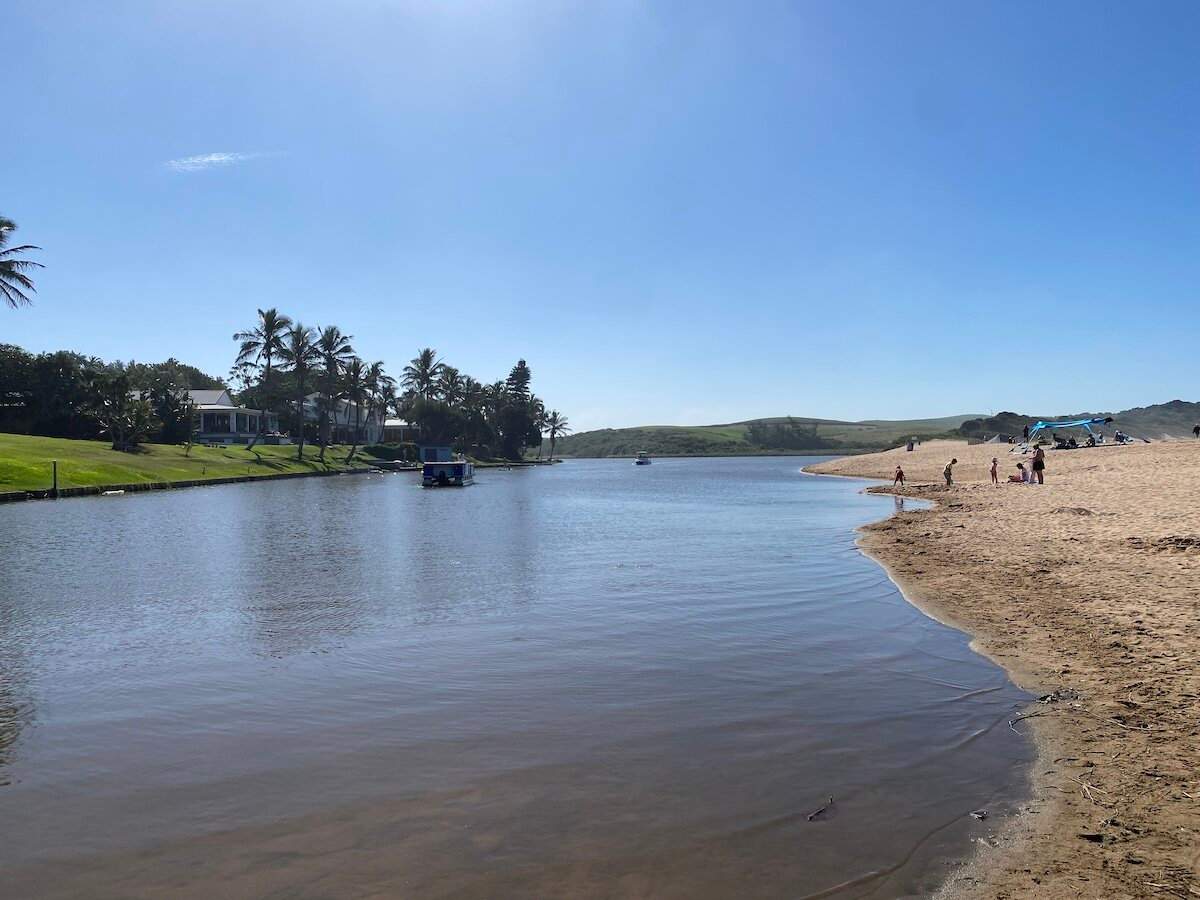 People enjoying the lagoon and sandy beach in Zinkwazi, with palm trees and distant hills under a clear sky.