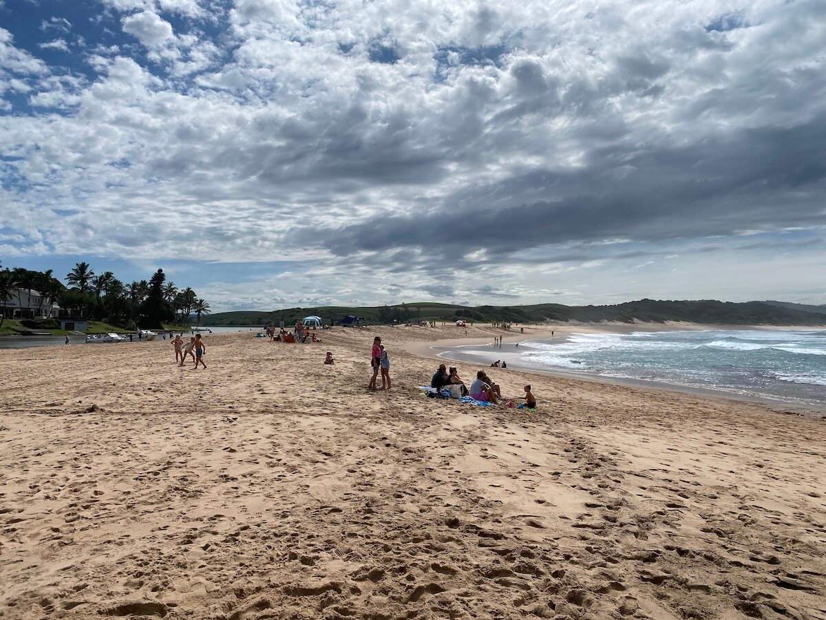 Families relax on the expansive sandy beach with the ocean and lagoon under a cloudy sky at Zinkwazi.