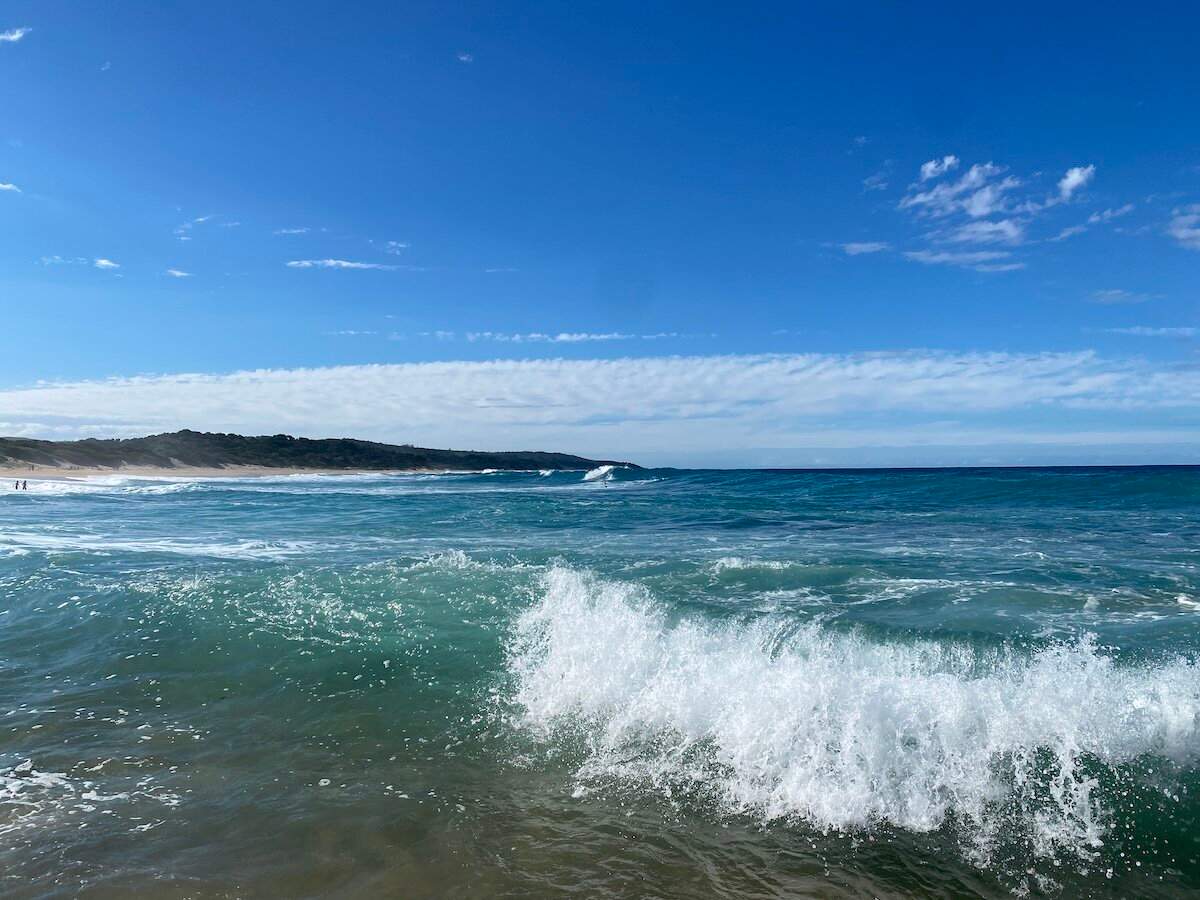 Waves crash near the sandy beach and lush coastline of Zinkwazi under a blue sky with scattered clouds.