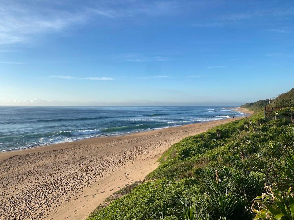 Expansive sandy beach with ocean waves under a clear blue sky, seen from a lush green hillside in Zinkwazi.
