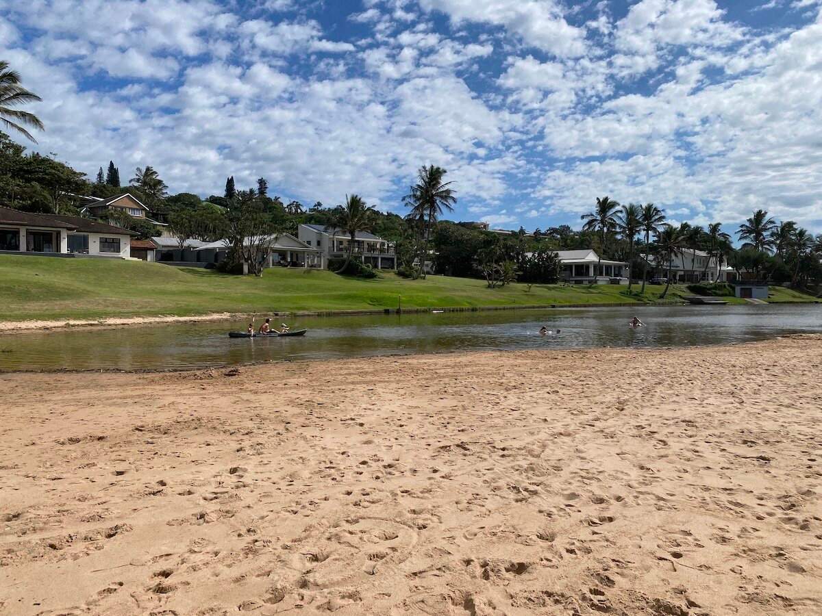 Sandy beach leading to a tranquil lagoon with kayakers, swimmers, and homes along the bank in Zinkwazi.