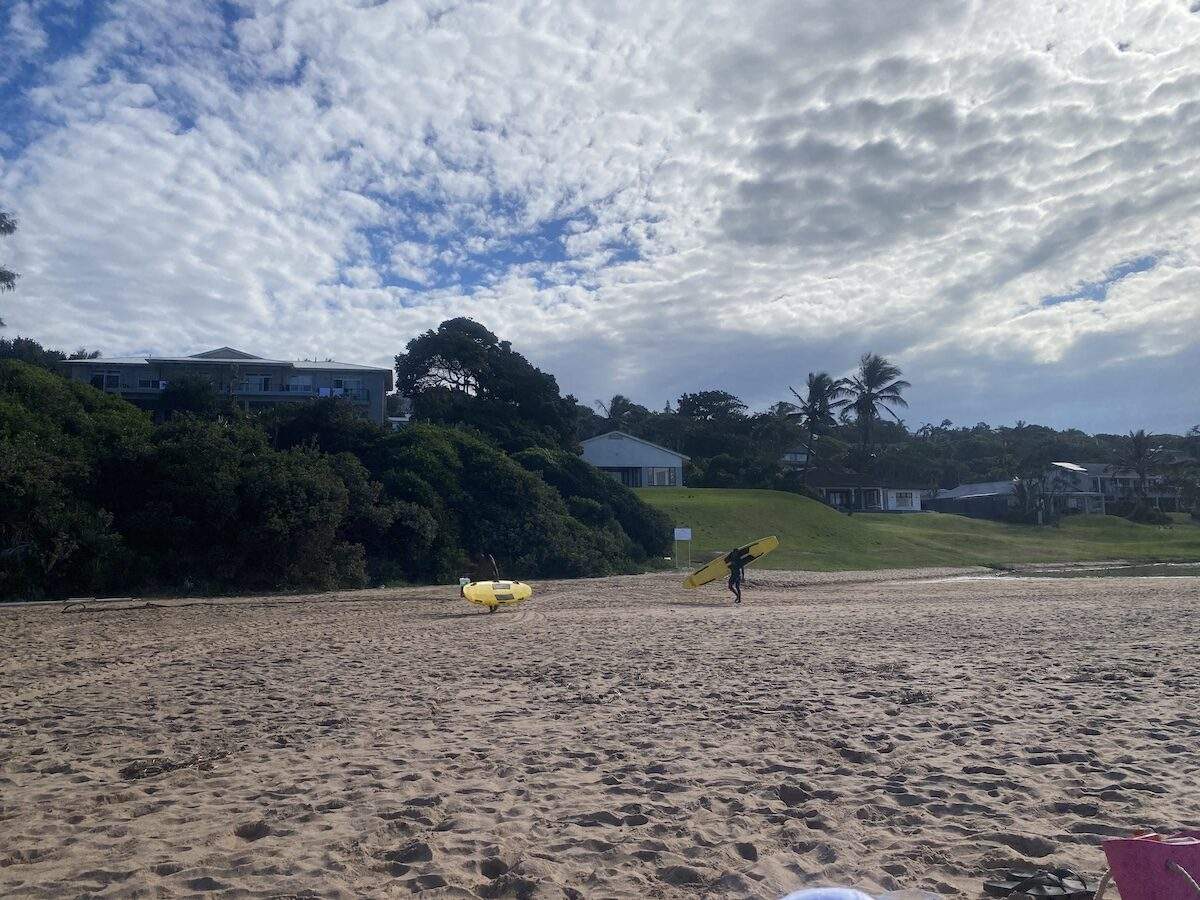 People with a surfboard and beach toys on a sandy beach at Zinkwazi with houses nestled in lush greenery.