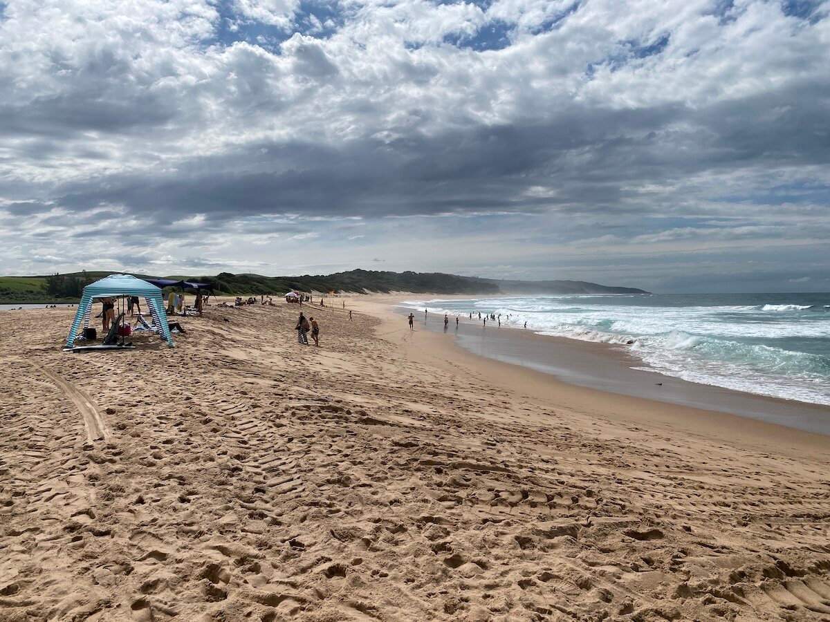 Cloudy day at Zinkwazi beach with people enjoying the ocean and sandy shore under a blue striped canopy.
